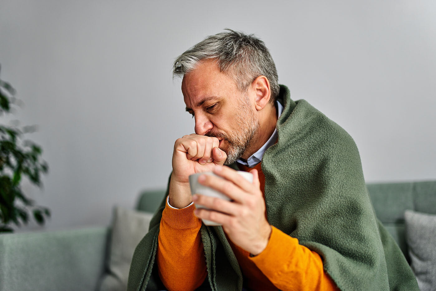 Mann, der mit Grippe erkrankt zu Hause auf dem Sofa sitzt und sich die Nase putzt
