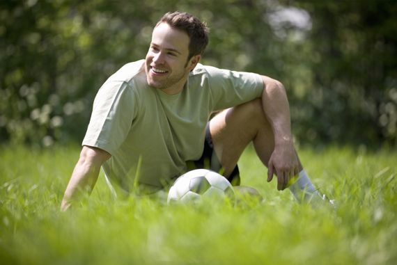 Mann in Sportkleidung sitzt mit Fußball auf Rasen